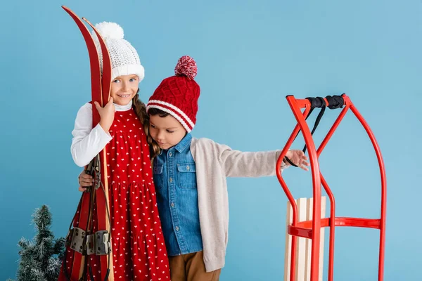 Ragazza in abito invernale in possesso di sci e abbracci fratello in piedi vicino slitta isolato su blu — Foto stock