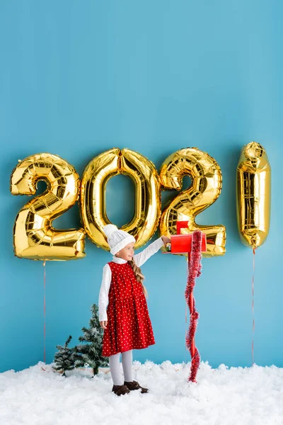 Girl in winter outfit taking present from mailbox near balloons with numbers while standing on snow on blue — Stock Photo