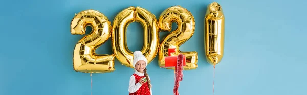 Панорамна концепція дівчини в зимовому вбранні, що тримає подарунок біля поштової скриньки і повітряних кульок з цифрами на синьому — стокове фото