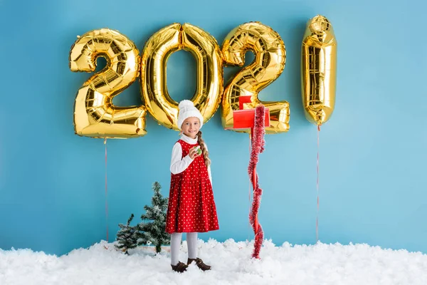 Kid in winter outfit holding present near mailbox and balloons with numbers while standing on snow on blue — Stock Photo