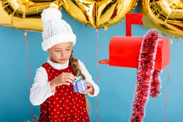 Дівчина в зимовому вбранні тримає подарунок біля червоної поштової скриньки на синьому — стокове фото