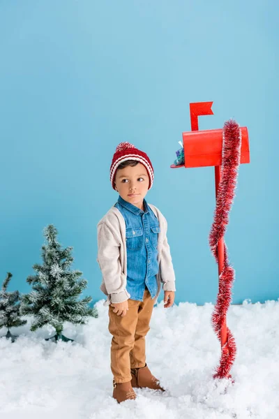 Garçon en chapeau et tenue d'hiver debout près de la boîte aux lettres rouge avec présent sur bleu — Photo de stock