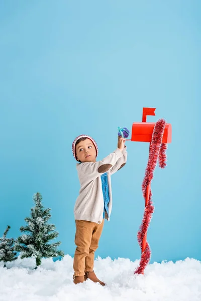 Мальчик в шляпе и зимнем наряде стоит рядом с красным почтовым ящиком, принимая подарок на синий — стоковое фото
