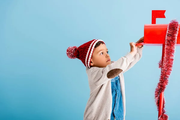 Boy in hat and winter outfit reaching red mailbox on blue — Stock Photo