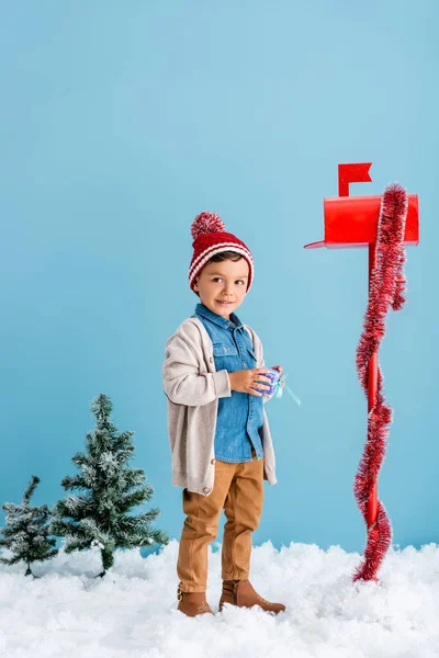 Хлопчик в зимовому вбранні тримає подарунок біля червоної поштової скриньки, стоячи на синьому — стокове фото