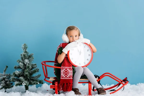Girl in winter earmuffs and scarf sitting in sleigh and holding clock near pines on blue — Stock Photo