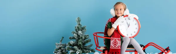 Panoramic concept of girl in winter earmuffs and scarf sitting in sleigh and holding clock near pines on blue — Stock Photo