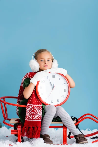 Girl in winter earmuffs and scarf sitting in sleigh and holding clock on blue — Stock Photo