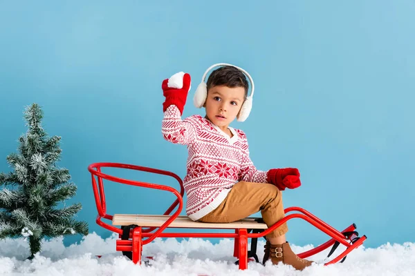 Boy in winter earmuffs and sweater sitting in sleigh and holding snowball on blue — Stock Photo