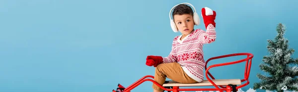 Horizontal image of boy in winter earmuffs and sweater sitting in sleigh and holding snowball isolated on blue — Stock Photo