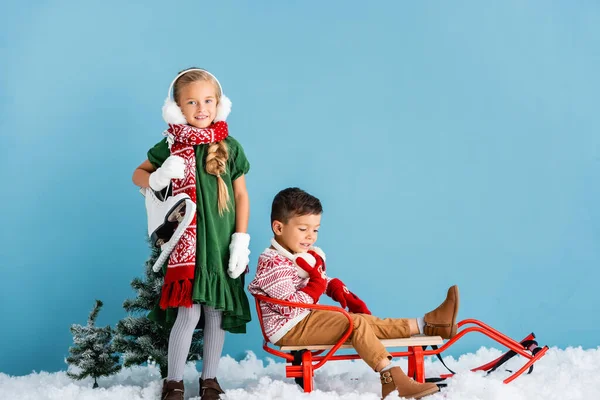 Girl in winter earmuffs and scarf standing with ice skates near boy in sleigh on blue — Stock Photo