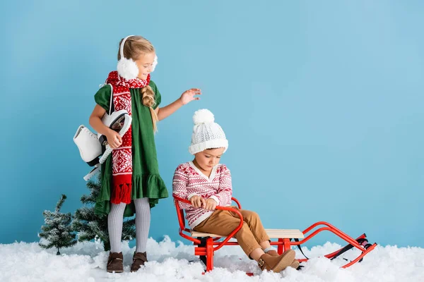 Mädchen mit winterlichen Ohrenschützern und Schal stehen mit Schlittschuhen und schauen den Jungen im Schlitten auf blau an — Stockfoto