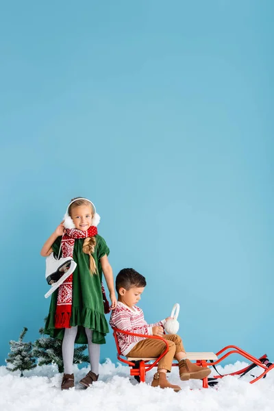 Enfant en hiver cache-oreilles et écharpe debout avec des patins à glace près du garçon en traîneau sur bleu — Photo de stock