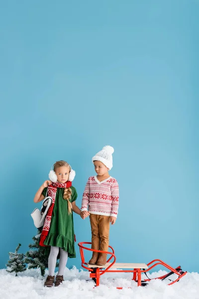 Fille en hiver cache-oreilles et écharpe debout avec des patins à glace et tenant la main avec frère dans le chapeau près du traîneau sur bleu — Photo de stock