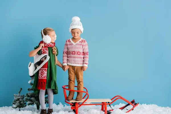 Girl in winter earmuffs standing with ice skates and holding hands with brother in hat near sleigh on blue — Stock Photo