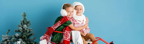 Panoramic crop of girl in winter earmuffs sitting on sleigh and hugging brother in hat on blue — Stock Photo