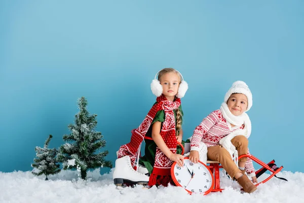 Kinder in Schals und Winterbekleidung sitzen auf Schlitten in der Nähe Uhr, Kiefern und Schlittschuhe auf blau — Stockfoto