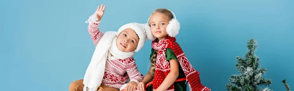 Panoramic concept of boy waving hand near girl in winter outfit sitting near pine on blue — Stock Photo