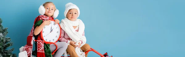 Horizontale Ernte von Kindern im Winteroutfit und Schals sitzen auf Schlitten mit Uhr in der Nähe von Kiefern auf blau — Stockfoto