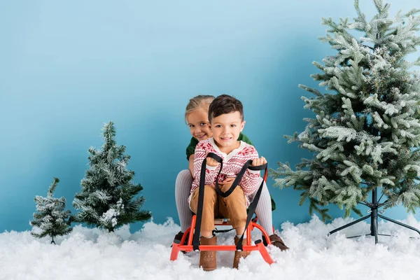 Kids sitting in sleigh on white snow near green pines on blue — Stock Photo