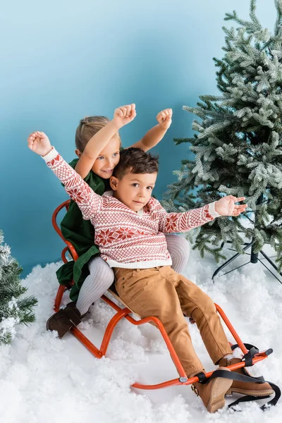 Crianças excitadas com as mãos acima das cabeças sentadas no trenó na neve branca perto de pinheiros verdes no azul — Fotografia de Stock