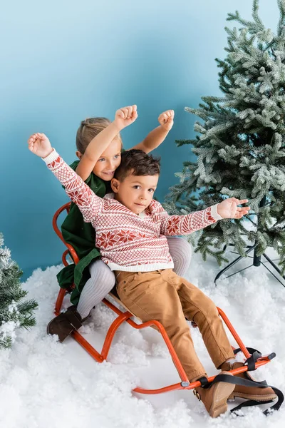 Excited children with hands above heads sitting in sleigh on white snow near green pines on blue — Stock Photo