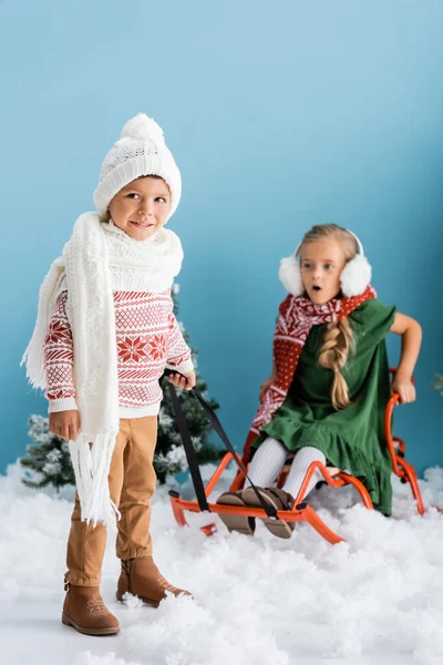 Selective focus of boy in scarf and hat giving a ride to sister on sleigh on blue — Stock Photo