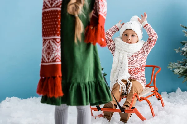 Selective focus of boy in winter hat sitting with hands above head on sleigh near sister on blue — Stock Photo