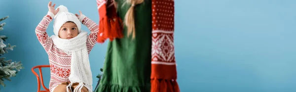 Panoramic crop of boy in winter hat sitting with hands above head near sister on blue — Stock Photo