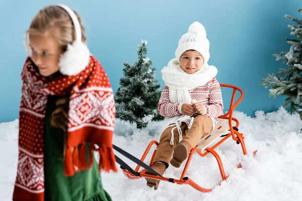 Selective focus of boy in winter hat sitting on sleigh near sister on blue — Stock Photo