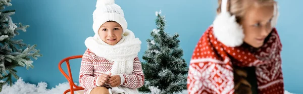 Horizontal crop of boy in winter hat sitting on sleigh near sister on blue — Stock Photo