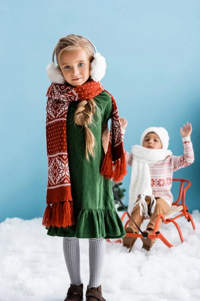 Selective focus of girl in winter outfit giving a ride to boy in hat on sleigh on blue — Stock Photo