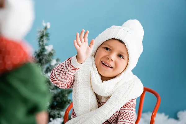 Selective focus of boy in knitted scarf and hat waving hand on blue — Stock Photo