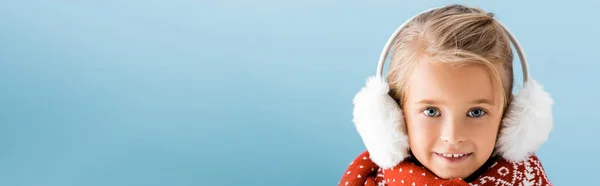 Panoramic crop of kid in winter earmuffs looking at camera isolated on blue — Stock Photo