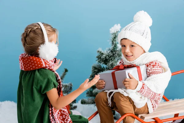 Junge mit Strickmütze hält Geschenk in der Hand, während er auf einem Taschenspielertrick in der Nähe von Mädchen in Winterohrenschützern sitzt, die auf blau gestikulieren — Stockfoto