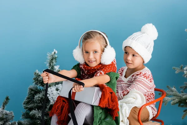 Kids in winter outfit riding sleigh with present near pines on blue — Stock Photo