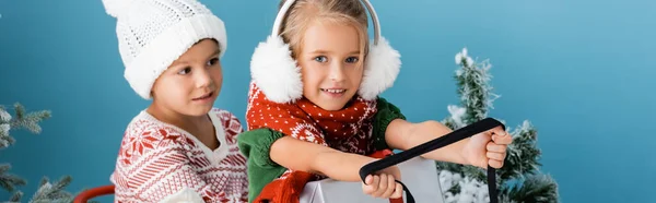 Panoramic crop of kids in winter outfit riding sleigh with present near pines on blue — Stock Photo