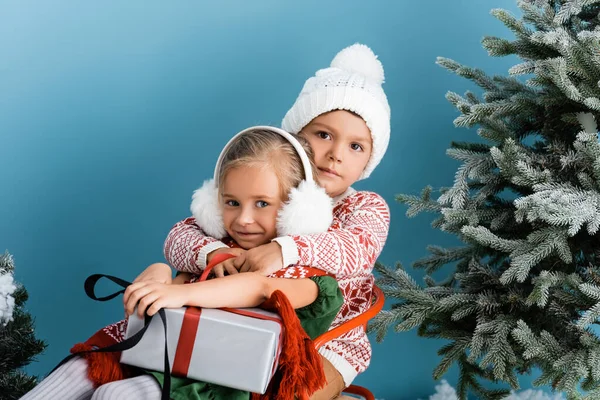 Брат в шляпе обнимает сестру, сидя у сосен с подарком на голубом — стоковое фото
