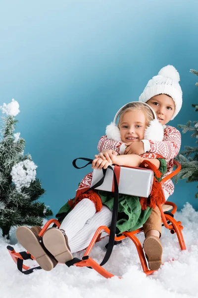 Junge mit Hut umarmt Schwester, während er in der Nähe von Kiefern mit Geschenk auf blau sitzt — Stockfoto