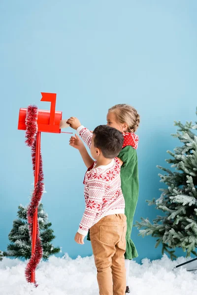 Kids reaching envelope in red mailbox with decoration near pines on blue — Stock Photo