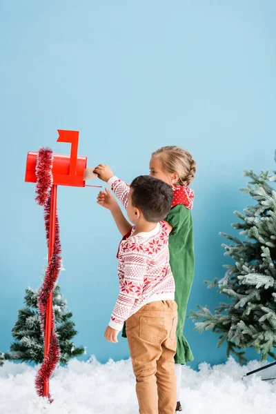 Niños alcanzando sobre en buzón rojo con decoración cerca de pinos en azul - foto de stock