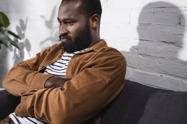 Selective focus of african american man sitting on couch with crossed arms — Stock Photo