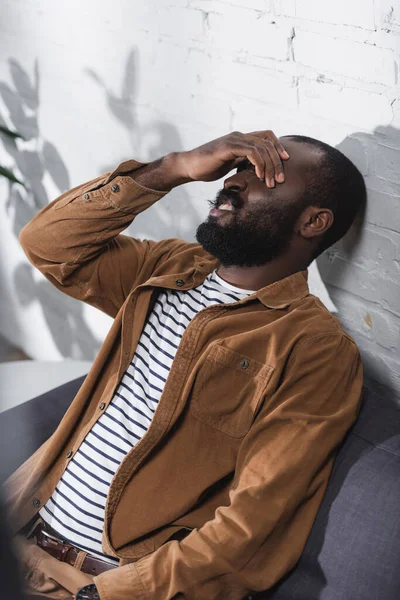 Enfoque selectivo del hombre afroamericano tocando los ojos y sentado en el sofá - foto de stock