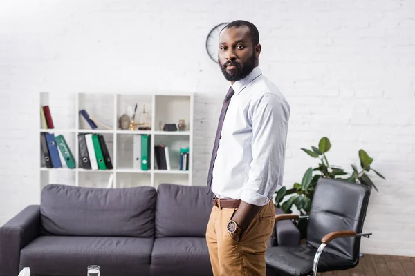 Bärtiger afrikanisch-amerikanischer Arzt in offizieller Kleidung, der in die Kamera blickt, während er mit den Händen in den Taschen im Krankenhaus steht — Stockfoto