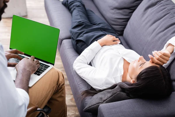 Enfoque selectivo de psicólogo afroamericano utilizando el ordenador portátil con pantalla verde cerca de la mujer acostada en el sofá - foto de stock