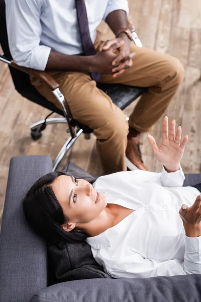 Вид зверху пацієнта розмовляє і жестикулює, лежачи на дивані біля афроамериканського психолога — стокове фото