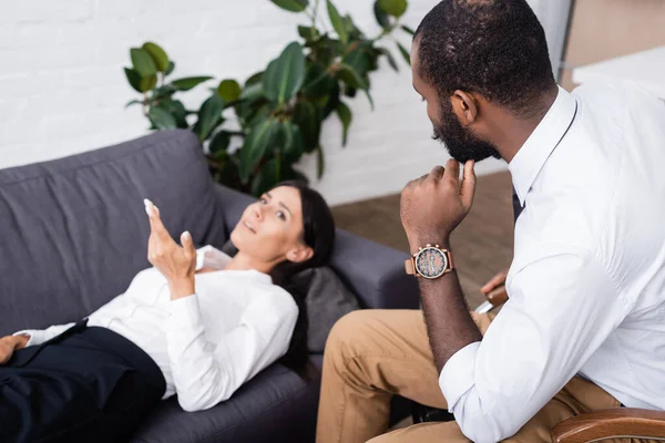 Enfoque selectivo de la mujer hablando y haciendo gestos mientras está acostado en el sofá cerca de psicólogo afroamericano - foto de stock