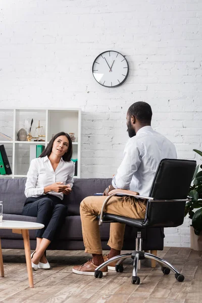 Thoughtful woman looking away while sitting on sofa and talking to african american psychologist — Stock Photo