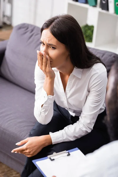 Selektiver Fokus einer aufgebrachten Frau, die Mund und Hand bedeckt, während sie in der Nähe eines Psychologen im Krankenhaus sitzt — Stockfoto