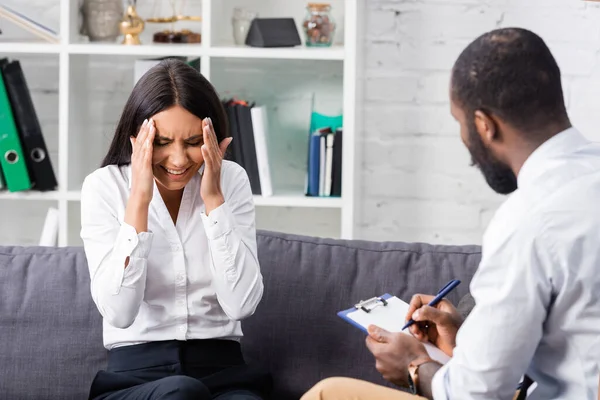 Messa a fuoco selettiva della donna stressata toccare la testa mentre seduto vicino a psicologo americano africano scrivere prescrizione sugli appunti — Foto stock